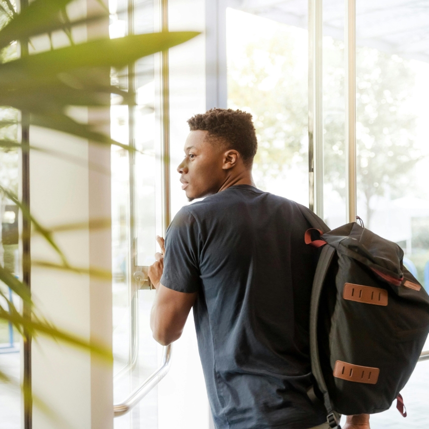 Man carrying black backpack with leather accents over one shoulder, near a glass door.