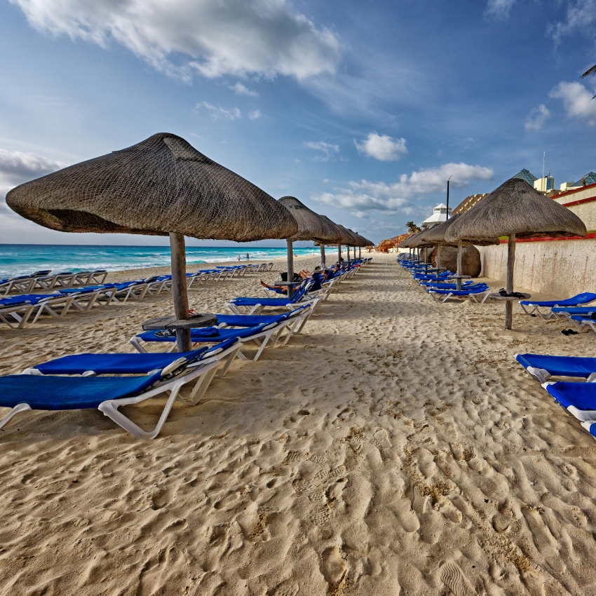 Beach with rows of blue lounge chairs under straw umbrellas.