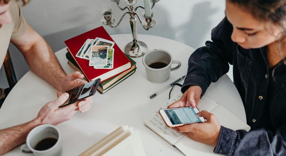 People using smartphones at a white table; notebooks, books, playing cards, and coffee mugs are also visible.