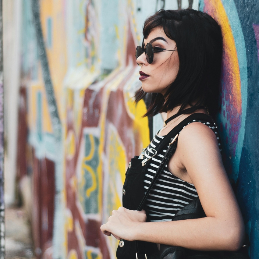Woman wearing round sunglasses, black-striped top, and black dungarees, leaning against a graffiti wall.