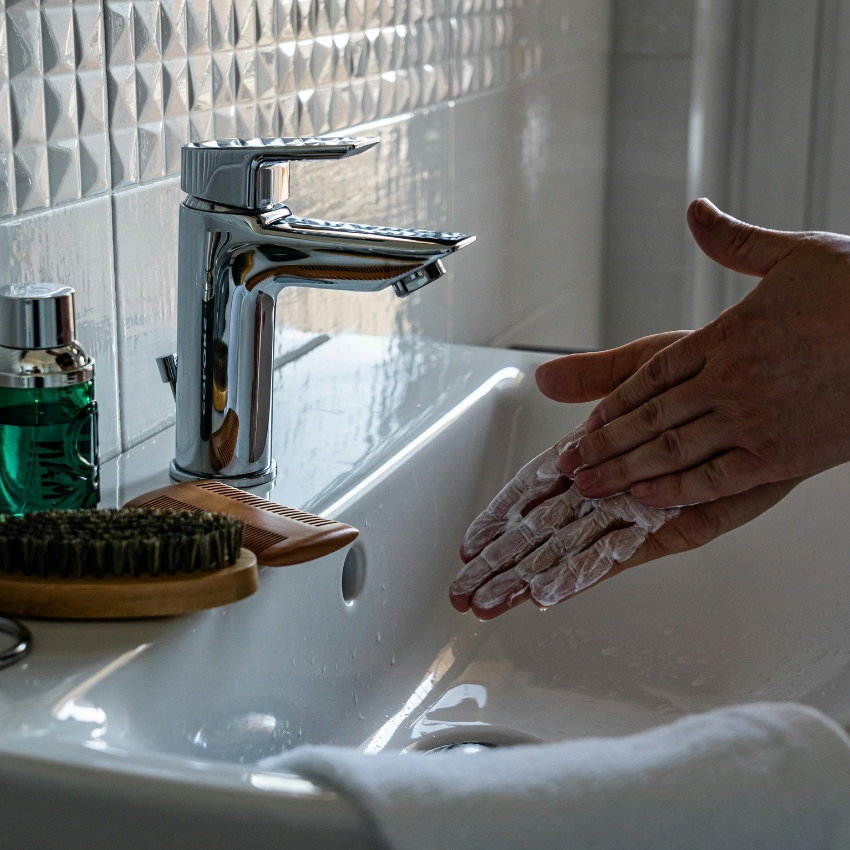 Hands washing with soap at a modern chrome faucet beside a grooming brush and green liquid soap bottle.