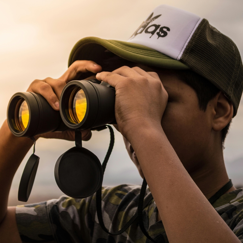 Person looking through black binoculars with yellow-tinted lenses; wearing a white and green cap.