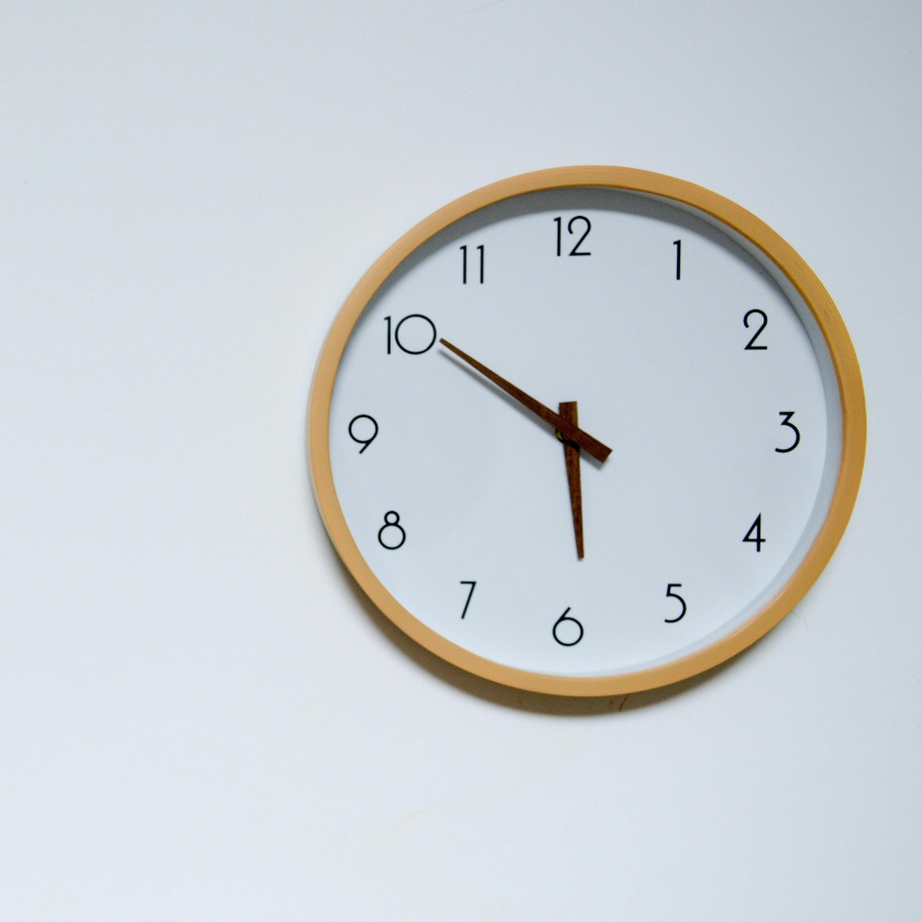 Wall clock with a light wood frame, white face, and black numbers. Time reads 10:12.