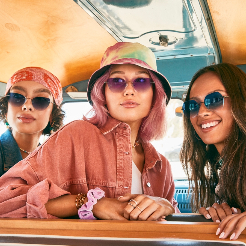 Three women wearing sunglasses; central woman with pink hair, two others with colorful headscarves.