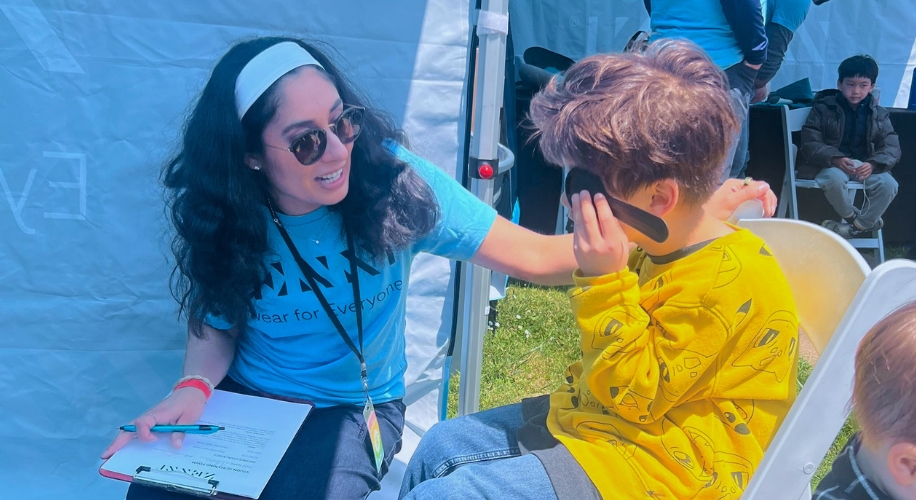 Child holds eye test occluder during vision screening with smiling caregiver.