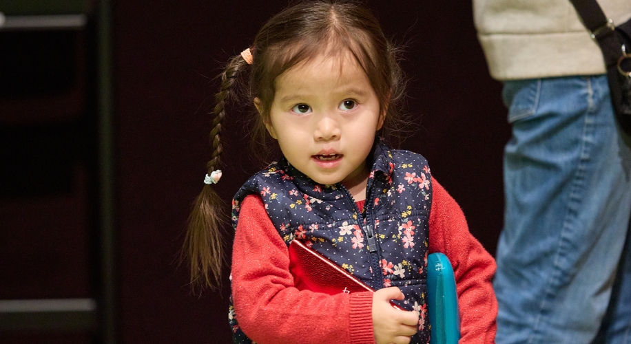 Young child with braided hair holding a blue object, wearing a floral vest and red sweater.