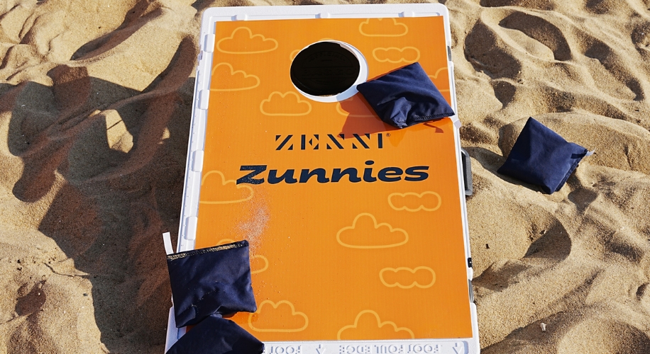 A bright orange cornhole board with navy bean bags sits on golden beach sand.