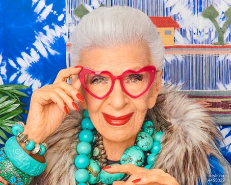 Iris Apfel wearing Iris Apfel x Zenni heart-shaped frame #4453027, along with bold turquoise jewelry, against a bright blue background.