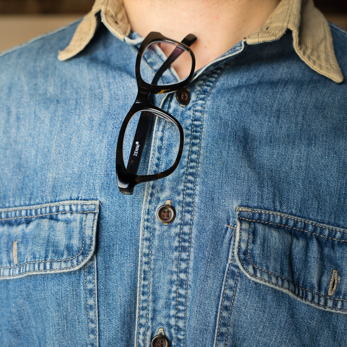 Black Maybeck eyeglasses from the Zenni Metropolitan Collection hanging from neck of chambray shirt.